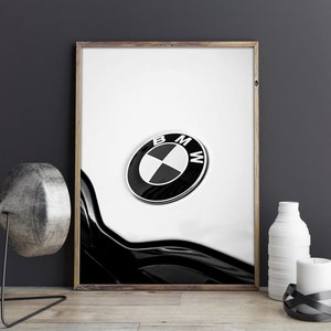 May include: Framed black and white print featuring the BMW logo. The logo is a black circle with a white and black quadrant design and the letters "BMW". The print is displayed on a wooden surface.