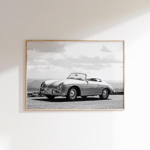 May include: Framed monochrome print featuring a classic silver convertible car. The car is parked on a paved surface with a mountain and cloudy sky backdrop. The car has a sleek design, evoking a sense of vintage elegance.