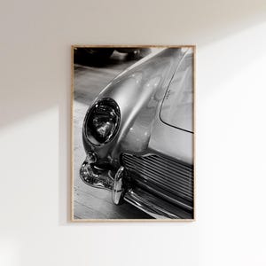 May include: Black and white framed print of a classic car's front end. The image highlights the headlight, chrome bumper, and grille of the vintage vehicle. The car's body has a shiny, reflective surface.