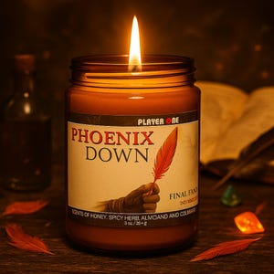 May include: A lit, amber-colored candle in a glass jar with the text "PHOENIX DOWN" and "PLAYER ONE" on the label. The label also features a red feather graphic and the text "FINAL FAN". The candle is 3 oz/25+g.