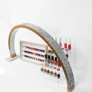 Puede incluir: Un arco dorado cubierto de pedrería, con una luz blanca en su interior, se encuentra sobre una superficie blanca. A la izquierda del arco hay una vitrina de madera con filas de botellas de esmalte de uñas en varios tonos de rojo y rosa. Delante del arco hay un expositor de acrílico transparente con filas de botellas de esmalte de uñas en varios tonos de rojo, rosa y marrón.