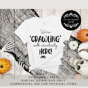 May include: White t-shirt with black text that reads "We're 'CRAWLING' with creativity Here!" and a black spider graphic. The shirt is lying on a wooden surface with black netting, a white skull, and orange and white pumpkins.