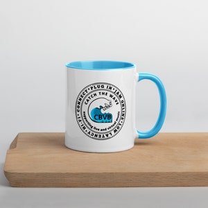 May include: White ceramic mug with a light blue interior and handle. The mug features a circular design with the text "CONNECT PLUG IN JAM ONLINE" and "CATCH THE WAVE" around a wave graphic. The text "CBVB" is also included.