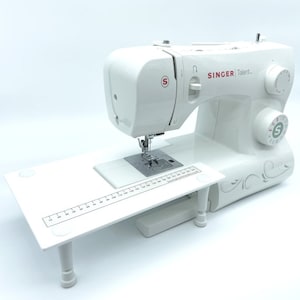 May include: White Singer Talent sewing machine with a white extension table and a ruler. The machine has a built-in needle threader and a stitch selection dial.