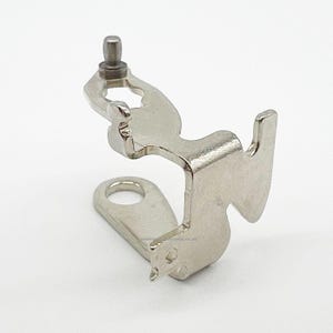 May include: A silver-coloured metal sewing machine component. It has a curved design with a small peg at the top and a circular opening at the bottom. The part has a unique shape with a hook-like extension.