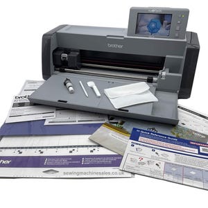 May include: A grey Brother ScanNCut DX die-cutting machine with a touchscreen display. The machine is open and shows a white cutting mat, a white tool holder, and a white tool. The machine is surrounded by various instruction manuals and a warranty card.