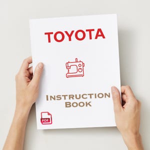 May include: A white instruction book held by two hands. The cover features the red word "TOYOTA" above a red line drawing of a sewing machine. Below the image is the text "INSTRUCTION BOOK" in brown.