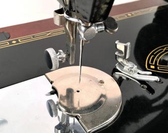May include: Close-up of a vintage black sewing machine. The image highlights the needle, presser foot, and other metallic components. The machine has a decorative gold emblem and a brown and gold accent on the top.