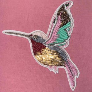 May include: A colourful hummingbird applique featuring gold, pink, green and red sequins. The applique is ideal for adding a touch of whimsy to clothing, bags or home decor.