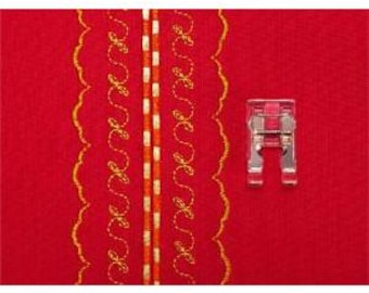 May include: A sewing machine presser foot next to a piece of red fabric with gold and orange decorative stitching. The presser foot is silver with a clear plastic component.