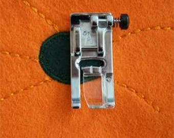 May include: A silver sewing machine foot with a clear plastic presser foot is positioned on an orange felt surface. A dark green felt circle is visible beneath the foot, with yellow stitching around it.