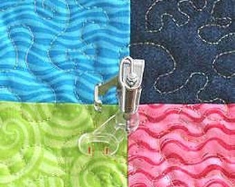May include: A clear quilting foot attached to a sewing machine, positioned over a patchwork quilt. The quilt features four squares in blue, green, pink, and navy, each with a different quilted pattern. The foot is made of clear plastic and metal.