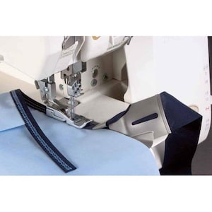 May include: A white overlocker sewing machine with a silver needle and presser foot. Blue fabric is being fed through the machine, with a finished seam in black and blue thread visible. The machine is on a light blue surface.