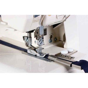 May include: A close-up of a white sewing machine with a needle and dark blue fabric being fed through. The machine has silver metal parts and a control panel. Ideal for sewing and fabric crafts.