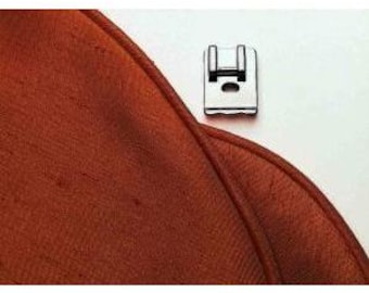 May include: A silver sewing machine presser foot. The foot is rectangular with a curved bottom and a hole in the center. It is positioned on a white surface next to a piece of rust-colored fabric.