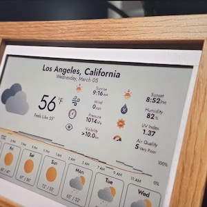 May include: A digital weather display framed in light wood, showing the weather forecast for Los Angeles, California. The screen displays the temperature at 56°F, cloud icons, wind speed, and a forecast for the next few days.