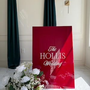 Puede incluir: Un gran letrero rojo brillante con el texto "The HOLLIS Wedding" en elegante escritura blanca. Un ramo de rosas blancas y paniculata se encuentra frente al letrero. Cortinas verde oscuro y un aplique de pared en el fondo.