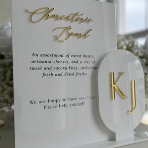May include: A white acrylic sign with gold lettering that reads "Charcuterie Board." The sign describes an assortment of cured meats, cheeses, and fruits. A smaller, attached piece displays the initials "KJ" in gold.
