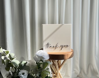 Könnte beinhalten: Ein weißes quadratisches Schild mit dem Text "thank you" in schwarzer Kalligraphie auf einem Holzhocker mit einer weißen Blume und grünen Blättern im Vordergrund.