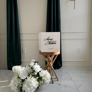 Puede incluir: Una caja blanca con los nombres "Annie & Nicholas" en escritura elegante, colocada sobre un pequeño taburete de madera. Un ramo de flores blancas está en el suelo, con cortinas verde oscuro al fondo.