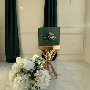 Peut inclure: Une boîte vert foncé avec l'inscription dorée "Mr. & Mrs. Colly" est posée sur un petit tabouret en bois. Un bouquet de fleurs blanches est devant. Des rideaux vert foncé sont en arrière-plan.