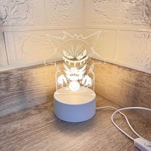 Puede incluir: Lámpara acrílica blanca iluminada, con un diseño de monstruo de dibujos animados. La lámpara se encuentra sobre una base redonda azul claro con el nombre "Alice" impreso. Un cable de alimentación blanco sale de la base.