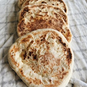 May include: A stack of four round, flatbreads with golden-brown toasted spots and a soft, pale interior. Each piece of bread has an irregular, circular shape with slightly charred edges, arranged in a vertical line on a textured fabric surface.