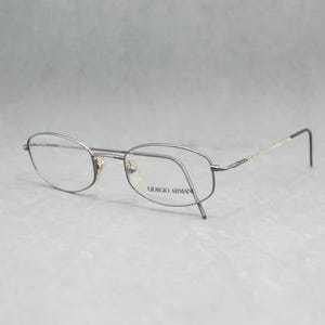May include: Silver-toned Giorgio Armani eyeglasses with oval lenses. The frames have a thin, delicate design with a subtle pattern on the temples. The glasses are set against a light gray background.