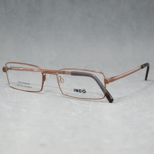Op de afbeelding: Rechthoekige bril met een lichtbruin metalen montuur en donkerbruine pootjes. De lenzen zijn helder en de merknaam "INDO" is zichtbaar op de rechterlens. Het montuur heeft ook de tekst "70% TITANIUM Light Non-allergenic."