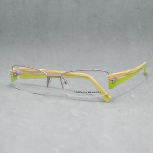 May include: Carolina Herrera spectacles with light brown and lime green striped temples. The frames are silver-coloured with a half-rim design. The brand name is printed on the lens.