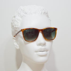 May include: A pair of brown tortoise shell sunglasses with green lenses. The sunglasses are on a white mannequin head.