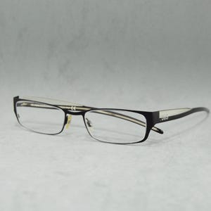 Peut inclure: Lunettes rectangulaires noires avec une fine bande blanche sur le dessus de la monture. Le nom de la marque "D&G" est visible sur la branche droite.