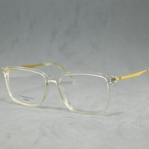 Puede incluir: Gafas de plástico transparente con patillas de metal dorado. Las gafas tienen una montura cuadrada.