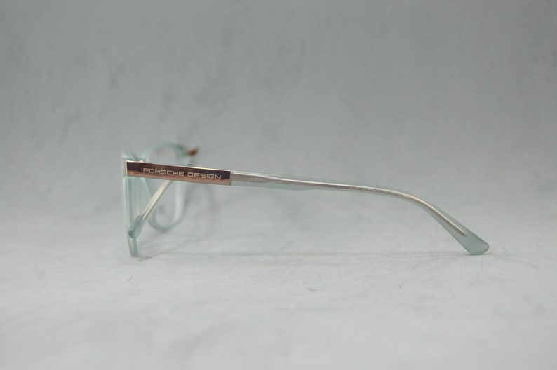 May include: A pair of light blue eyeglasses with a silver frame and rose gold accents. The frame has the text "PORSCHE DESIGN" on the side.