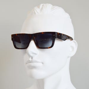 Puede incluir: Gafas de sol rectangulares con montura de carey en tonos marr&oacute;n y negro y lentes oscuras degradadas. La montura presenta un puente grueso y patillas anchas con un peque&ntilde;o detalle met&aacute;lico cerca de las bisagras, sobre un maniqu&iacute; blanco.