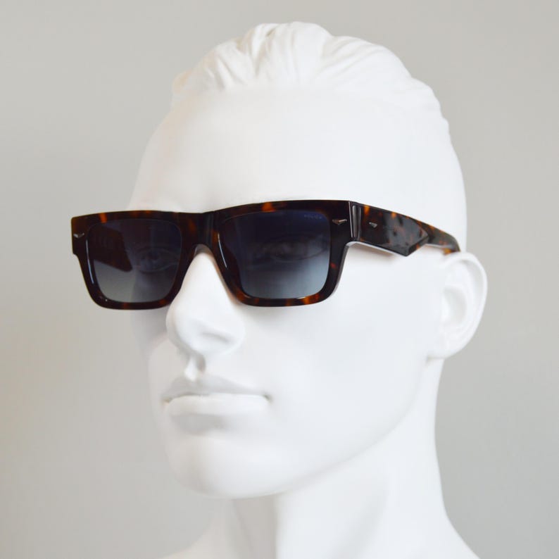 Puede incluir: Gafas de sol rectangulares con montura de carey en tonos marr&oacute;n y negro y lentes oscuras degradadas. La montura presenta un puente grueso y patillas anchas con un peque&ntilde;o detalle met&aacute;lico cerca de las bisagras, sobre un maniqu&iacute; blanco.