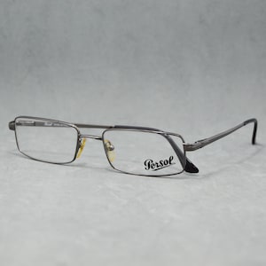 May include: Rectangular eyeglasses with a dark gray metal frame. The lenses are clear, and the brand name "Persol" is printed on the right lens. The temples are thin and curve down at the ends.