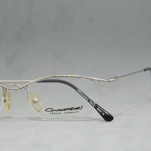 May include: Silver-toned metal eyeglasses with a black frame and clear lenses. The glasses have a decorative pattern on the top of the frame and the words "Conquistador Berlin Germany" printed on the right lens.