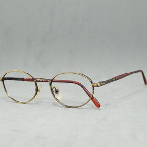 Può includere: Occhiali ovali con montatura color borgogna e bordo dorato.