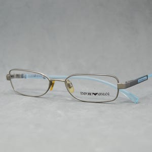 May include: Silver-framed eyeglasses with light blue temples. The rectangular lenses are set in a silver frame, with the brand name "EMPORIO ARMANI" printed on the right lens. The temples are a light blue color.