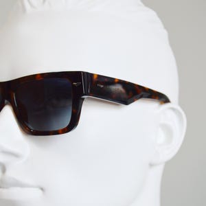 Puede incluir: Gafas de sol de carey con una montura rectangular gruesa y lentes oscuras degradadas. Las monturas presentan un distintivo patr&oacute;n moteado en marr&oacute;n y negro con un acabado pulido y peque&ntilde;os detalles en tono plateado en las bisagras de las patillas.