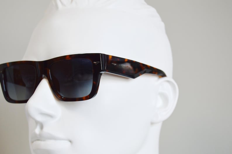 Puede incluir: Gafas de sol de carey con una montura rectangular gruesa y lentes oscuras degradadas. Las monturas presentan un distintivo patr&oacute;n moteado en marr&oacute;n y negro con un acabado pulido y peque&ntilde;os detalles en tono plateado en las bisagras de las patillas.