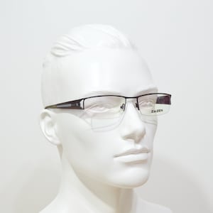 May include: A black half-rimmed spectacles frame with clear lenses. The frame has a silver accent on the top of the bridge. The brand name "Zazen" is printed on the right temple.
