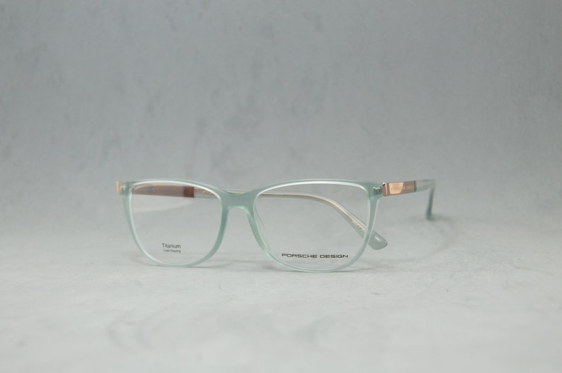 May include: A pair of light green spectacles with a gold accent on the arms. The frames are made of titanium and the lenses are clear. The text "Porsche Design" is printed on the right lens.