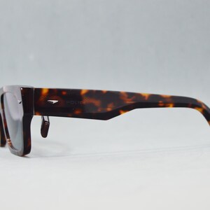 Puede incluir: Gafas de sol de carey con montura rectangular gruesa y lentes oscuras. Las patillas presentan un corte angular cerca de la bisagra y un peque&ntilde;o detalle met&aacute;lico plateado. La palabra Police aparece sutilmente grabada en el lateral.