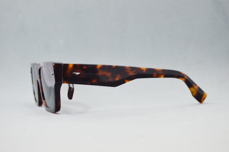 Puede incluir: Gafas de sol de carey con montura rectangular gruesa y lentes oscuras. Las patillas presentan un corte angular cerca de la bisagra y un peque&ntilde;o detalle met&aacute;lico plateado. La palabra Police aparece sutilmente grabada en el lateral.