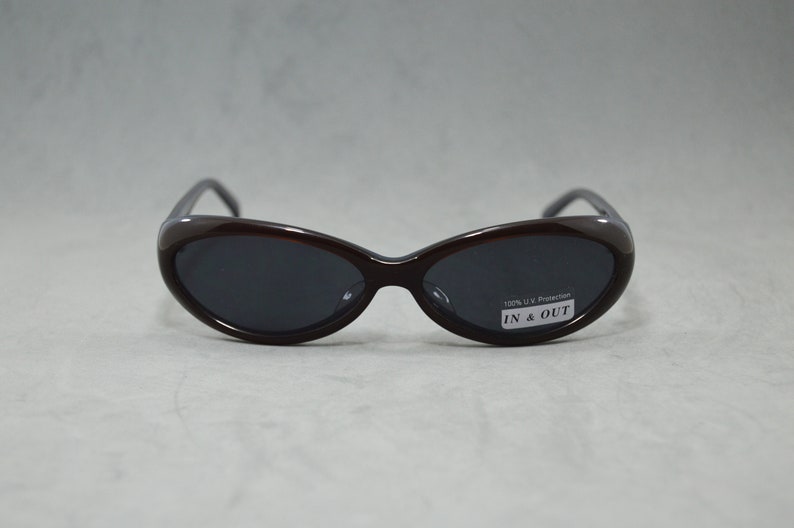 May include: Brown oval-shaped sunglasses with dark lenses. The sunglasses have a label that reads "100% UV Protection IN & OUT".
