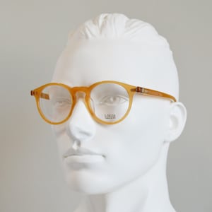 May include: Amber-colored round-frame glasses with a translucent finish. The frames feature a keyhole bridge and thin temples. The right lens is printed with the text LOZZA, followed by the smaller text "l'occhiale dal 1878" in black lettering.