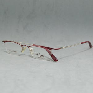 Può includere: Occhiali da vista con montatura in metallo rosso e oro con lenti trasparenti. Il nome del marchio "VOS" è stampato sulla lente destra.