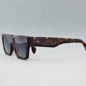 Puede incluir: Gafas de sol de carey con una montura gruesa y rectangular con patrones moteados en marr&oacute;n oscuro y &aacute;mbar. Las lentes son de color gris oscuro y las patillas son anchas, con un peque&ntilde;o detalle met&aacute;lico plateado cerca de la bisagra.